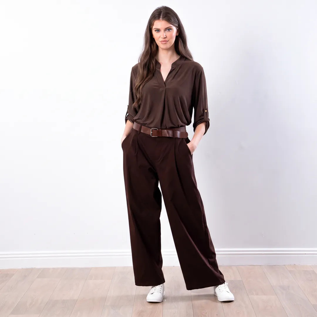 Sharlene Top in brown — relaxed jersey blouse with notch V neckline and roll-tab sleeves, styled with brown wide-leg trousers and white trainers, full-length front view