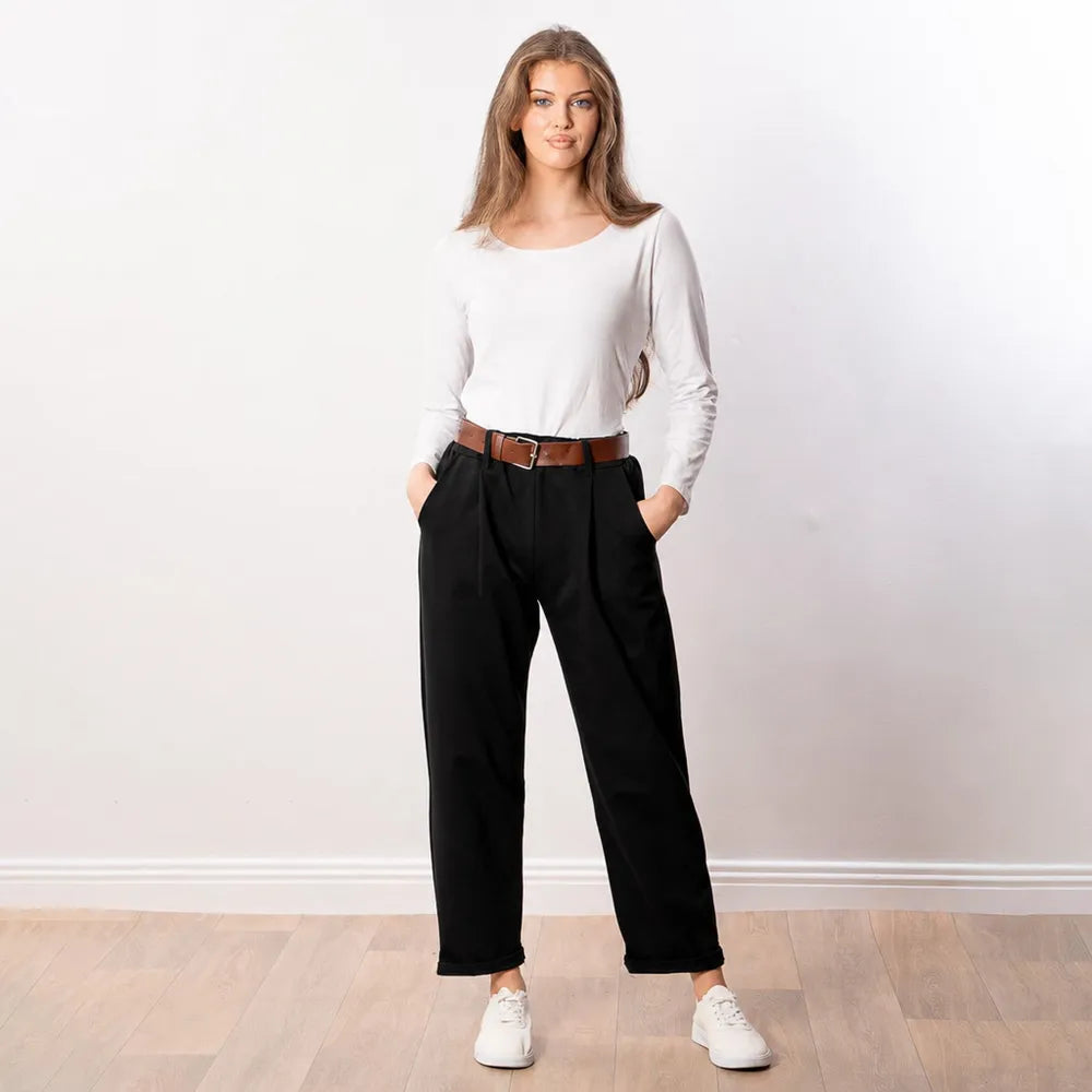 Woman wearing black Arya tapered trousers with a white long-sleeve top and tan leather belt, front view with hands in pockets